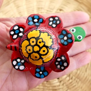 May include: A hand-painted, red flower-shaped ornament with yellow and black center, and blue and white accents. The ornament has a green, turtle-like appendage with white eyes. The design is colorful and detailed, showcasing a folk art style.