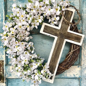May include: A white and brown wooden cross with a white painted frame is attached to a wreath of woven twigs and white blossoms. The wreath is hanging on a blue wooden door.