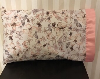 May include: A rectangular pillow with a butterfly pattern in shades of pink, gray, and brown on a white background. The pillow has a pink satin border. The pillow is resting on a black surface.