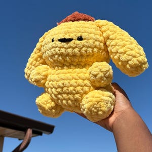 May include: A handmade, yellow crochet plush toy with a small, brown hat. The plush has black eyes and a black nose. The toy is held up against a bright blue sky.