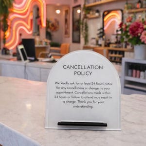 May include: A white acrylic sign displaying the "CANCELLATION POLICY" in a salon. The sign outlines the policy, with a black bar at the base. The background features a neon sign and shelves stocked with products, creating a professional atmosphere.
