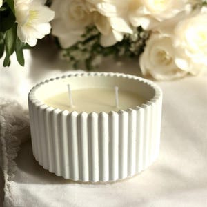 May include: A white, ribbed candle in a round container with two wicks. The candle is set on a white surface, with a blurred backdrop of white flowers. The candle's design features vertical ridges around the exterior.