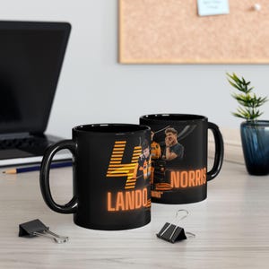 May include: Two black ceramic mugs with orange and white designs. One mug features the number "4" and the text "LANDO" in orange. The other mug features the text "NORRIS" in white. Both mugs have a black handle.