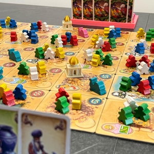 Peut inclure: Un plateau de jeu avec des pièces en bois colorées et un petit bâtiment blanc au centre. Le plateau est en carton et a un fond brun avec un design de carte. Il y a aussi plusieurs cartes en arrière-plan.