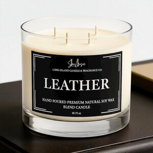 K&ouml;nnte beinhalten: Ein durchsichtiger Glas-Kerzenhalter mit cremefarbenem Wachs. Die Kerze hat drei Dochte und ein schwarzes Etikett mit wei&szlig;em Text, der "LEATHER" lautet. Das Etikett enth&auml;lt auch den Text "LONG ISLAND CANDLE & FRAGRANCE CO." und "35.75 oz."