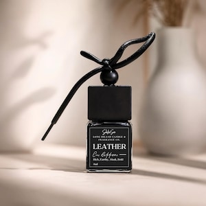 May include: A black car air freshener with a leather scent. The rectangular glass bottle has a black cap and a leather strap. The label reads "LEATHER Car Diffuser" and "Rich, Earthy, Musk, Bold."