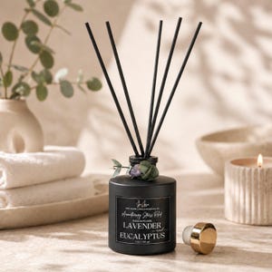 May include: A black reed diffuser with black reeds, a gold-colored cap, and a label that reads "Lavender and Eucalyptus." The diffuser is surrounded by a candle, towels, and a vase with greenery. The product is 7 oz / 197 ml.