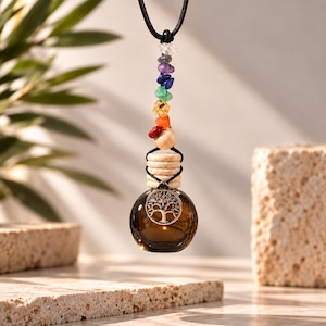 May include: A hanging car diffuser bottle made of dark brown glass, featuring a silver-toned tree of life charm on the front. The bottle is suspended by a black cord adorned with a series of small, multi-colored gemstone beads and a wooden bead.