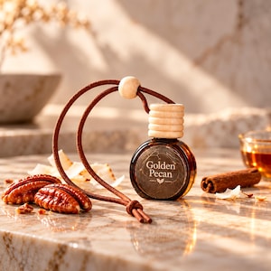 May include: A small, round glass bottle filled with amber liquid, featuring a label that reads Golden Pecan. The bottle has a light-colored wooden cap and a brown suede cord for hanging. It is surrounded by whole pecans and a cinnamon stick.