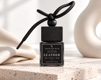 May include: A square glass car diffuser bottle with a black wooden cap and a black cord tied in a bow. The label reads Curated Long Island, Car Diffuser, Leather, Rich, Earthy, Musk, 0.33 fl oz, 10 ml. The bottle contains a dark liquid.
