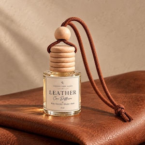 May include: A clear glass car diffuser bottle with a light wood cap and a round wooden bead. A brown cord is attached for hanging. The label reads: Curated Long Island, LEATHER Car Diffuser, Rich, Earthy, Musky, Bold, 8 ml. The bottle contains liquid.