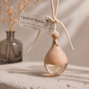 May include: A wooden car air freshener with a clear glass bottom, filled with a light-colored liquid. The freshener is suspended by a cream-colored cord and a tag that reads "Opulent Smoke" from Long Island Candle & Fragrance Co.