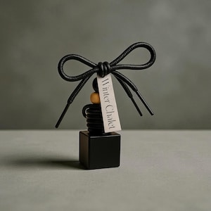 May include: A black car air freshener with a bow made of shoelaces. The air freshener has a black square base, a coiled black center, and a small tag that reads "Winter Chalet". The air freshener has a neutral color palette.