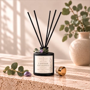 May include: A matte black glass reed diffuser bottle with five black reeds, featuring a label that reads Curated Long Island Lavender Eucalyptus Aromatherapy 7 fl oz | 207 ml. A gold-toned stopper and a purple amethyst stone sit nearby.