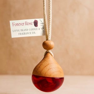 May include: A wooden and glass air freshener with a red liquid interior, suspended by a cream-colored rope. The top is a light wood color, and the bottom is a teardrop shape. A tag reads "Forever Rose" and "Long Island Candle & Fragrance Co."