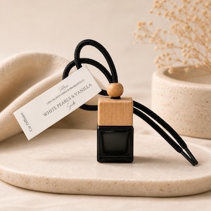 May include: A car air freshener with a black glass bottle and a wooden top. The label reads "White Pearls & Vanilla Suede." The freshener hangs from a black cord with a wooden bead. The product is displayed on a neutral-toned surface.
