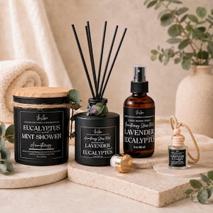 May include: Aromatherapy products including a candle, reed diffuser, linen room spray, and car diffuser. The candle is in a black tin with a wooden lid, labeled "Eucalyptus Mint Shower." The reed diffuser is black with reeds, labeled "Lavender Eucalyptus." The spray bottle is amber, labeled "Lavender Eucalyptus."
