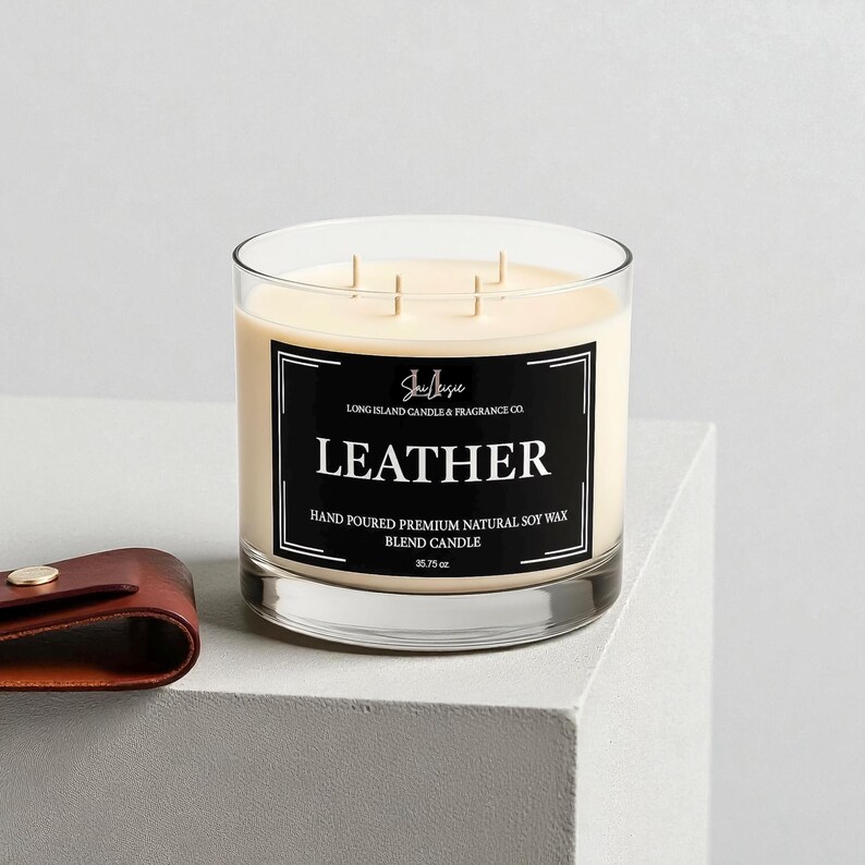 K&ouml;nnte beinhalten: Eine Glaskerze mit cremefarbenem Wachs und drei Dochten. Die Kerze hat ein schwarz-wei&szlig;es Etikett mit der Aufschrift "LEATHER" und "HAND POURED PREMIUM NATURAL SOY WAX BLEND CANDLE". Die Kerze wiegt 1013 Gramm.