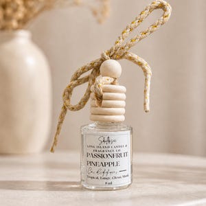 May include: A clear glass car air freshener with a wooden top and a decorative rope. The label reads "Passionfruit Pineapple Car Diffuser". The scent is described as tropical, tangy, citrus, and musk. The bottle contains 8 ml.