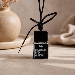 May include: A black car air freshener with a square bottle and a black string for hanging. The label reads "CEDARWOOD BLANC Car Diffuser" with the scent described as "Zesty, Earthy, Warm". The bottle contains 8 ml.