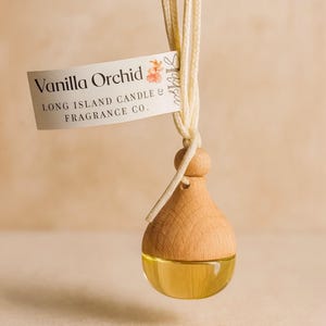 May include: A hanging air freshener with a wooden top and a clear glass bottom filled with yellow liquid. The label reads "Vanilla Orchid" and "Long Island Candle & Fragrance Co.". The air freshener is suspended by a cream-colored cord.