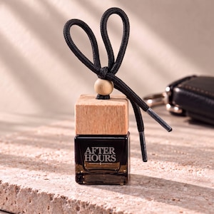 May include: A small square glass bottle with a dark tint and a light wood cap. The text AFTER HOURS is printed in white on the front. A black cord is tied in a loop at the top with a small wooden bead, featuring metal aglets at the ends.