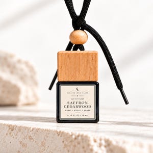 May include: A square glass car diffuser bottle with a light wood cap and a black cord featuring a wooden bead. The white label reads Curated Long Island, Car Diffuser, Saffron Cedarwood, Warm, Woody, Amber, 0.33 fl oz | 10 ml in black text.