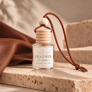 May include: A clear glass 8 ml car diffuser bottle with a light wood cap and a brown suede hanging cord. The white label features the text: Curated Long Island, LEATHER Car Diffuser, Rich, Earthy, Musky, Bold, 8 ml. The bottle is topped with a wooden bead.