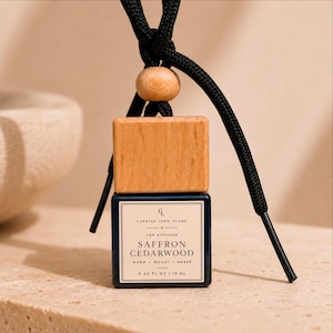 May include: A dark blue glass car diffuser bottle with a light wood cap and a black hanging cord. The label reads Curated Long Island, Car Diffuser, Saffron Cedarwood, Warm Woody Amber, 0.33 fl oz | 10 ml. A small wooden bead sits above the cap.