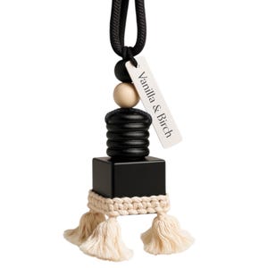 May include: Black and cream-colored car air freshener with a macrame design and tassels. The air freshener has a black square base, a wooden bead, and a tag that reads "Vanilla & Birch". The air freshener hangs from a black cord.