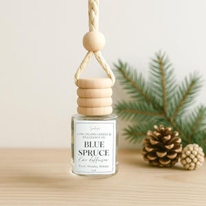 May include: A clear glass bottle with a wooden cap and a string for hanging. The bottle contains a car diffuser with the label "Blue Spruce Car Diffuser" and the text "Long Island Candle & Fragrance Co.". The bottle is sitting on a wooden surface with a pine branch and pine cones in the background.