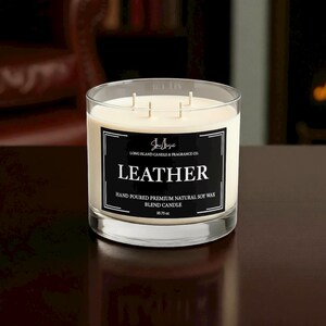 K&ouml;nnte beinhalten: Ein Glas-Kerze mit drei Dochten und cremefarbenem Wachs. Ein schwarzes Etikett zeigt "LEATHER" und "HAND POURED PREMIUM NATURAL SOY WAX BLEND CANDLE". Die Kerze wiegt 1013 Gramm.