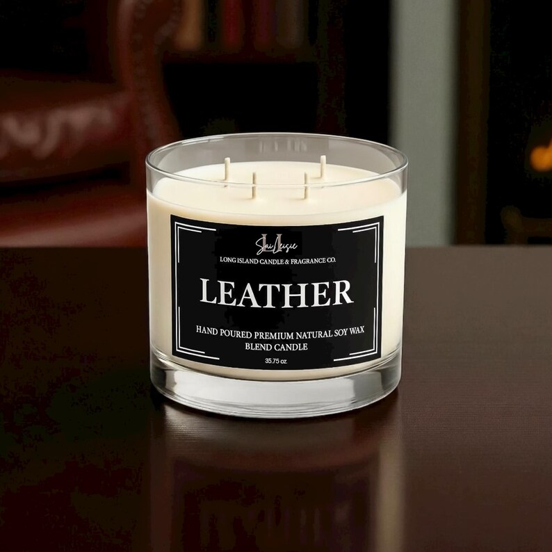 K&ouml;nnte beinhalten: Ein Glas-Kerze mit drei Dochten und cremefarbenem Wachs. Ein schwarzes Etikett zeigt "LEATHER" und "HAND POURED PREMIUM NATURAL SOY WAX BLEND CANDLE". Die Kerze wiegt 1013 Gramm.