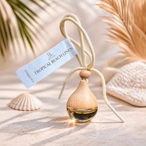 May include: A wooden car air freshener with a Tropical Beach Linen scent. The teardrop-shaped diffuser has a light wood top and a clear glass bottom filled with liquid. A tag with the text "TROPICAL BEACH LINEN" hangs from a cream-colored cord.