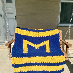 May include: A chunky knit blanket in navy blue and yellow, featuring a large yellow "M" in the center. The blanket has horizontal yellow stripes and is draped over a wooden swing.