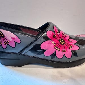 Puede incluir: Zueco gris con un gran diseño floral rosa. La flor tiene un centro amarillo y detalles negros. El zapato tiene una suela negra y un acabado brillante. El diseño está pintado a mano.