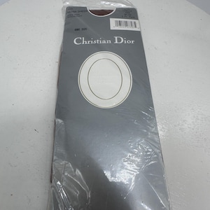 Peut inclure: Un paquet de collants Christian Dior Ultra Sheer de couleur Tan. L'emballage est blanc avec un ovale noir au centre. Le texte "Christian Dior" est imprimé en noir en haut de l'emballage. L'emballage est scellé avec un film plastique transparent.