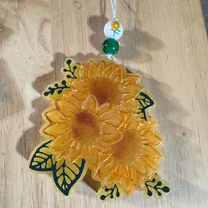 May include: A sunflower-shaped car air freshener with a white string, green and white beads, and a sunflower bead. The air freshener features three yellow sunflowers with green leaves and dark green outlines.