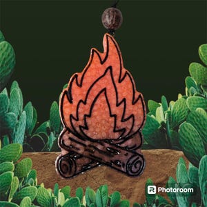 May include: A black and orange ceramic ornament shaped like a campfire with flames. The ornament has a textured surface and is hanging from a black cord.