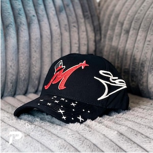 May include: A black baseball cap featuring a red rhinestone letter M topped with a small black and white top hat. The brim is decorated with white embroidered stars and dots, while the side displays a white embroidered signature-style graphic.