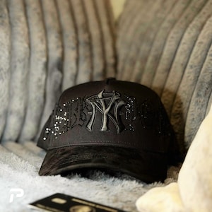 May include: A black baseball cap featuring a prominent embroidered NY logo on the front panel, accented with intricate black rhinestone detailing that extends across the sides. The brim is made of a textured black material with a subtle pattern.