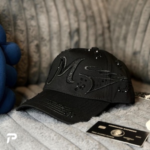 Puede incluir: Una gorra de béisbol negra con una letra M grande y estilizada adornada con cristales negros. La gorra incluye detalles adicionales de cristales negros dispersos por la corona y una visera curva con costuras. Se ve una tarjeta de crédito negra.