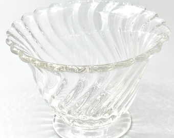 May include: A clear glass dessert bowl with a spiral design. The bowl has a scalloped rim and sits on a small, round base. The glass is transparent, allowing light to pass through.