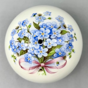 May include: A white ceramic salt or pepper shaker with a floral design. The design features a bouquet of blue forget-me-nots tied with a pink ribbon. The shaker has several small holes for dispensing the contents.
