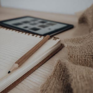 May include: A wooden pencil lies on a spiral-bound notebook with lined pages. An e-reader is in the background. A beige-coloured, woven textile is draped on the side. The scene is set on a light-coloured wooden surface.
