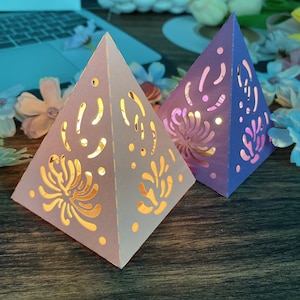 Może przedstawiać: Dwie papierowe latarnie w kształcie piramidy z misternymi wycięciami. Latarnia po lewej jest biała z różowym gradientem, a latarnia po prawej jest fioletowa z różowym gradientem.