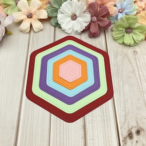 Può includere: Set di sei fustelle di carta a forma di esagono in vari colori, tra cui rosso, arancione, rosa, azzurro, viola e verde. Le fustelle sono sovrapposte, con la fustella più grande in basso e la più piccola in alto. Le fustelle hanno un bordo cucito.