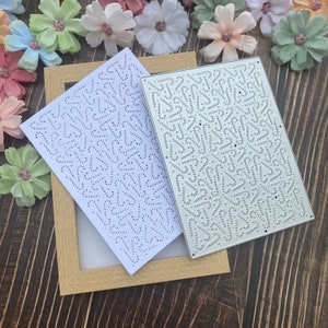 May include: Two metal craft dies with intricate patterns. One die is silver, the other is white. Both have a repeating design of small dots and curved lines. The dies are on a wooden surface, with a gold picture frame and artificial flowers.