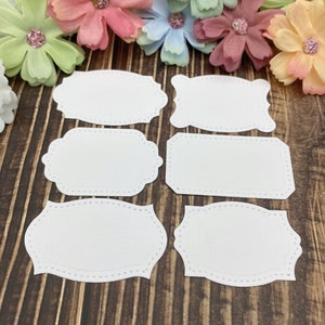 May include: Six white blank labels with different shapes and scalloped edges. The labels are on a brown wooden surface.