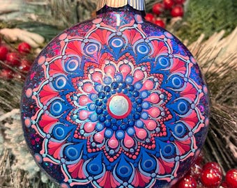 May include: A round, blue Christmas ornament with a detailed mandala design in shades of pink and blue. The ornament has a silver cap and is decorated with glitter. The mandala pattern is symmetrical and intricate, with a central silver circle.